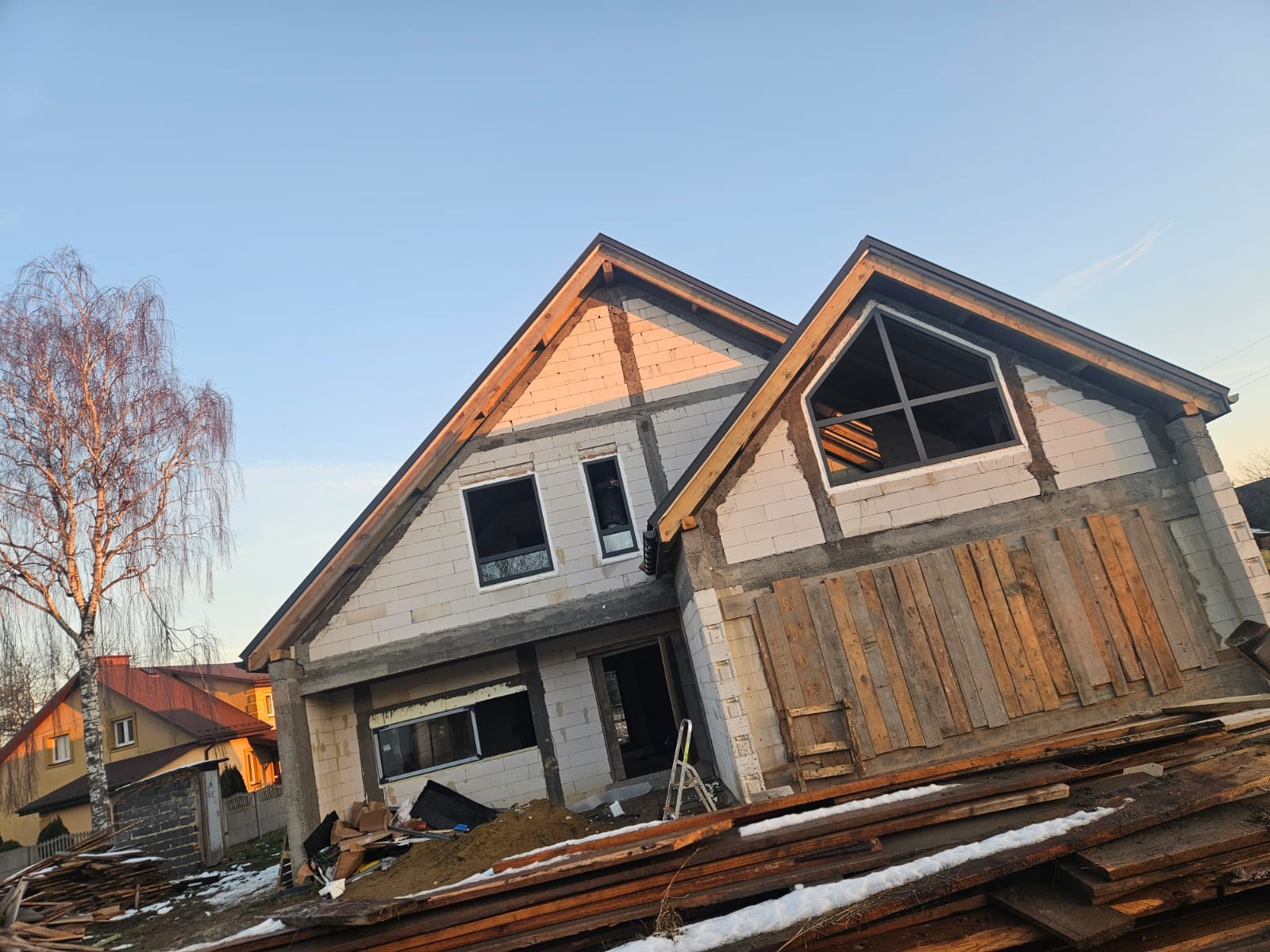 Dom w stanie surowym otwartym z zamontowanymi oknami aluminiowymi o nietypowym kształcie, drewniane deski zabezpieczające otwór, widoczne białe bloczki konstrukcyjne i przygotowane drewno na dachu.