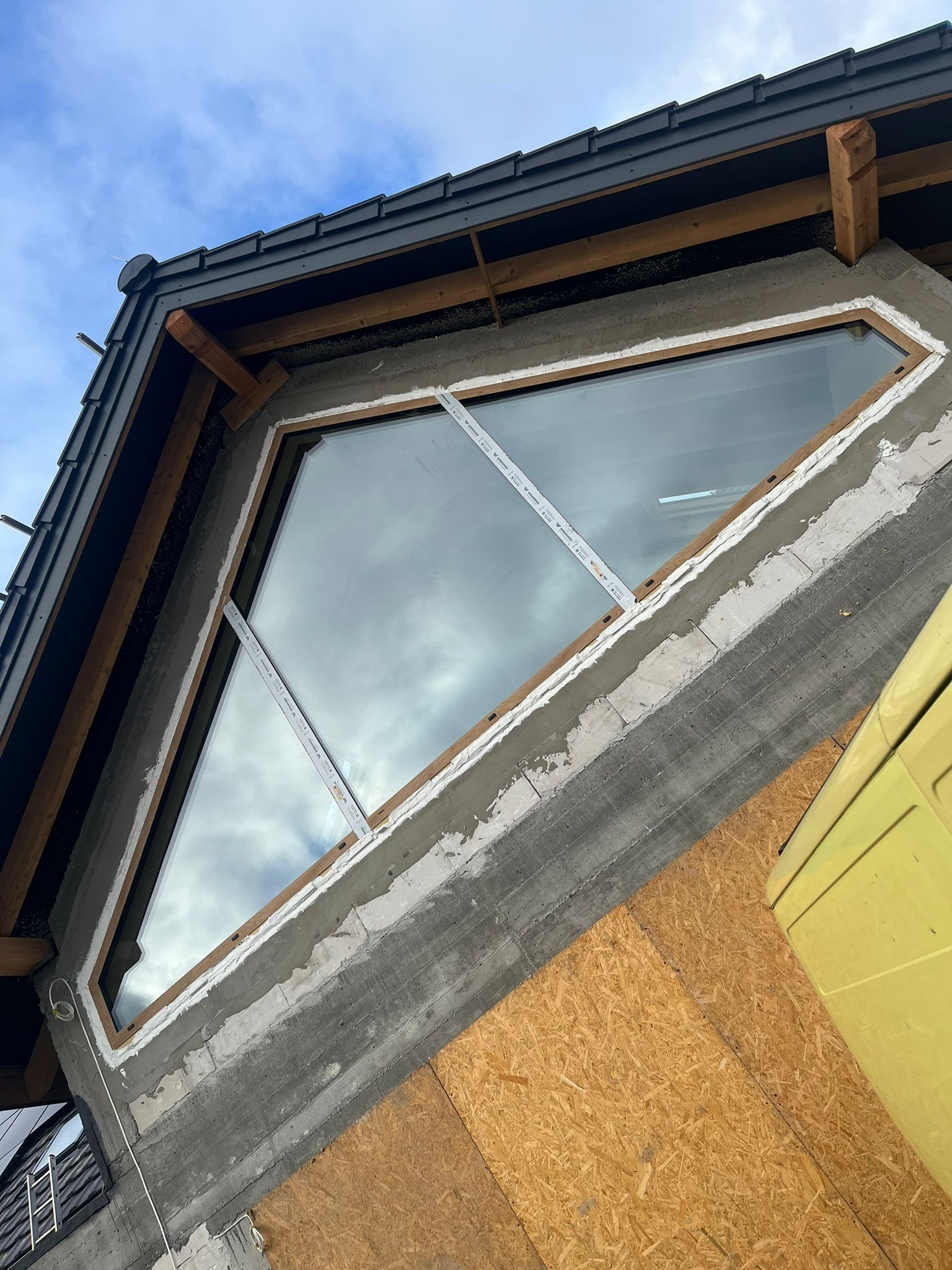 Trójkątne okno zamontowane w surowym stanie budynku, widoczne elementy konstrukcyjne dachu i fragment rusztowania.