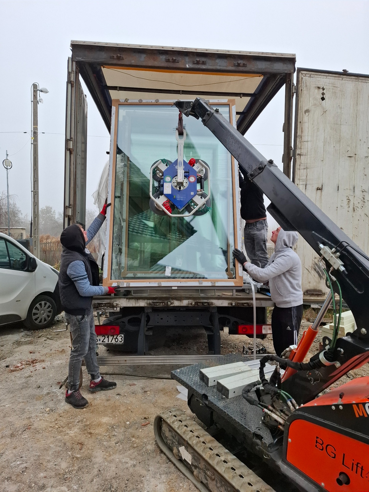 Montaż okna za pomocą przyssawki i dźwigu mini na pace samochodu dostawczego, dwóch monterów w trakcie pracy, widoczne detale mocowania i zabezpieczenia okna.