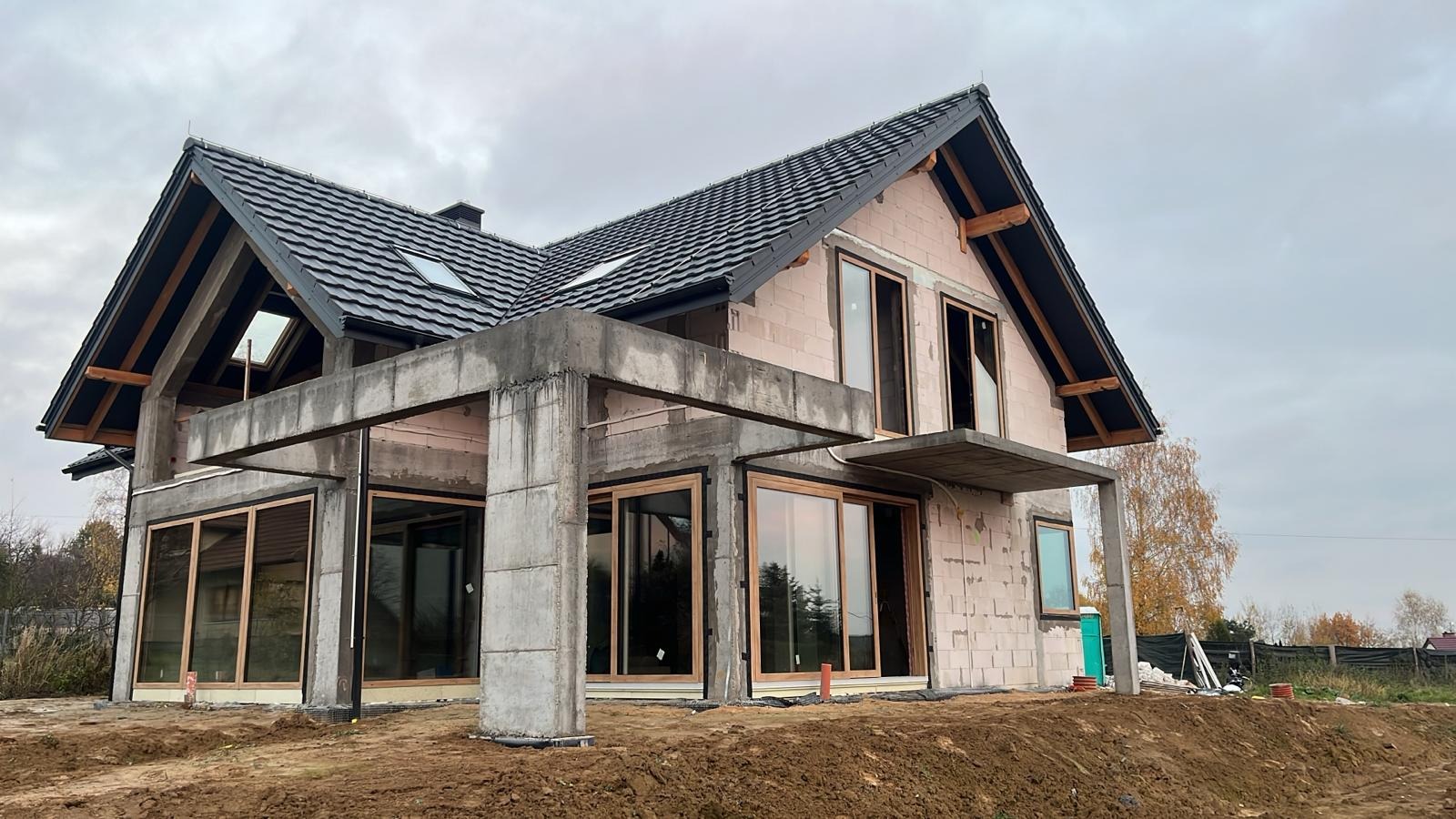 Dom w trakcie budowy z widocznymi wstawionymi oknami z drewnianymi ramami, betonową konstrukcją tarasu i czarnym dachem z dachówkami, otoczony ziemią i jesienną roślinnością.