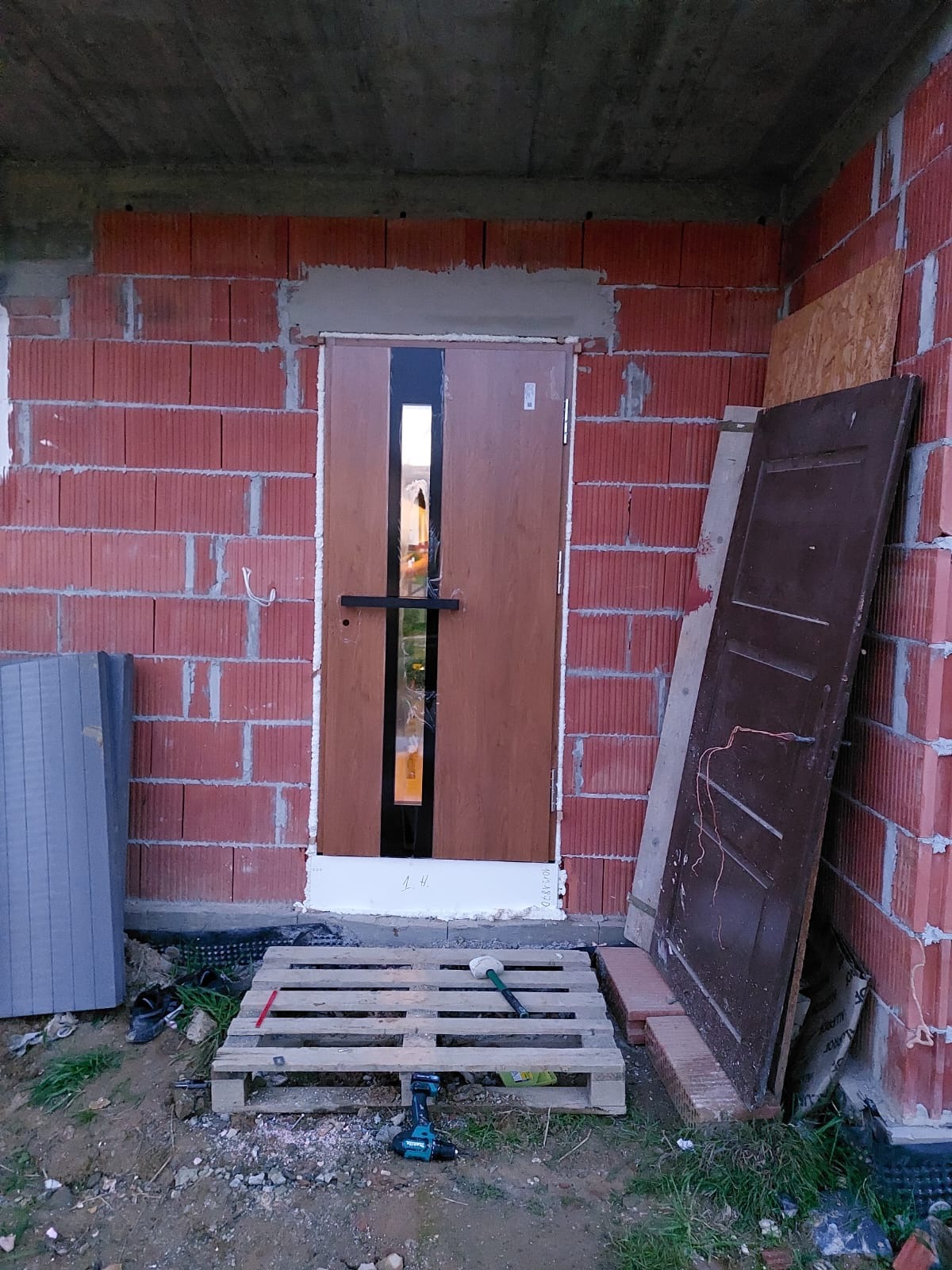 Montaż brązowych drzwi z czarnym paskiem w surowym stanie, widoczny mur z czerwonej cegły, paleta z narzędziami i stara, brązowa płyta drzwiowa oparta o ścianę.