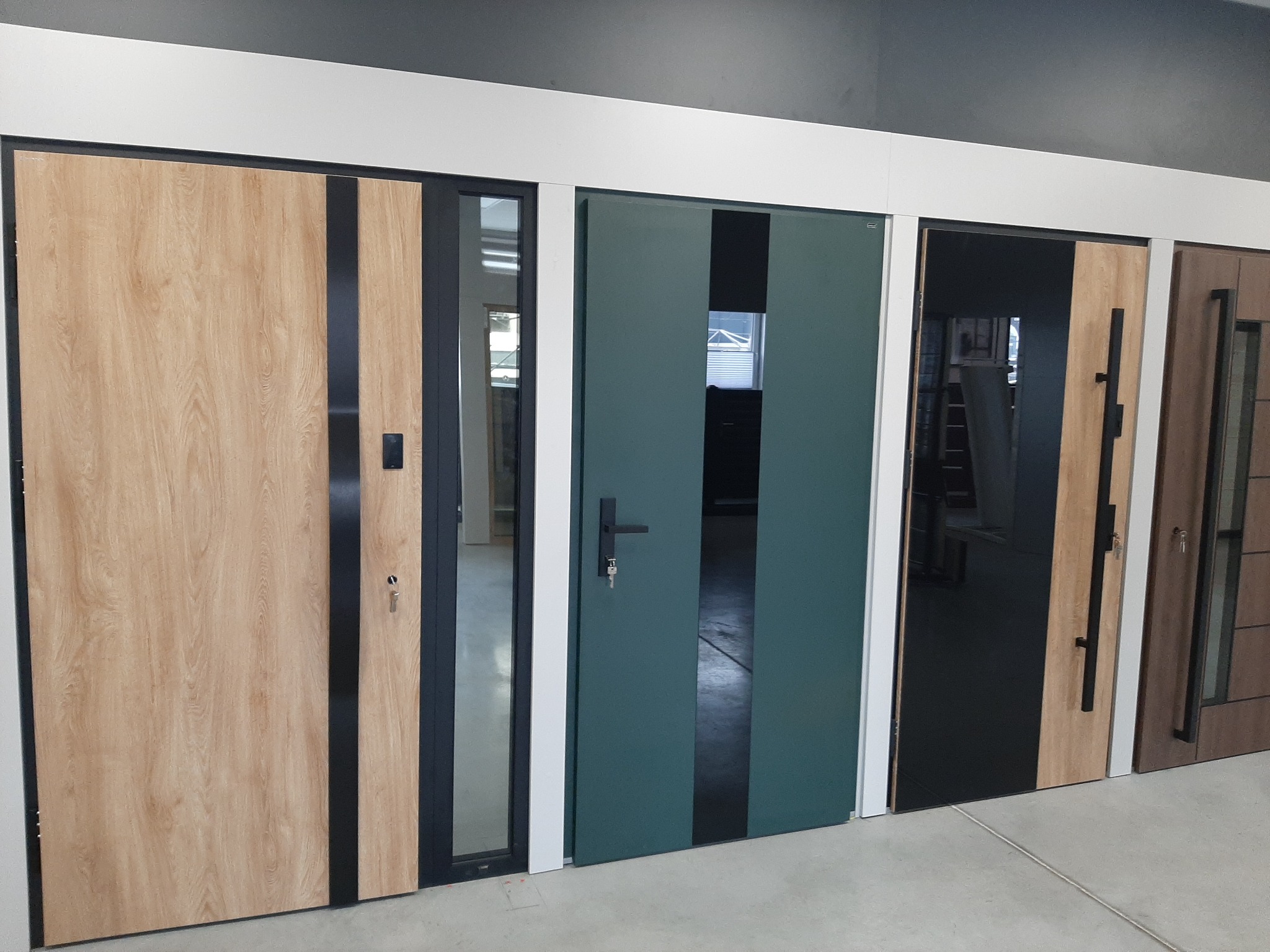 Ekspozycja czterech różnych modeli drzwi z pionowymi, czarnymi paskami dekoracyjnymi, w tym drzwi z przeszkleniami i w kolorze butelkowej zieleni.