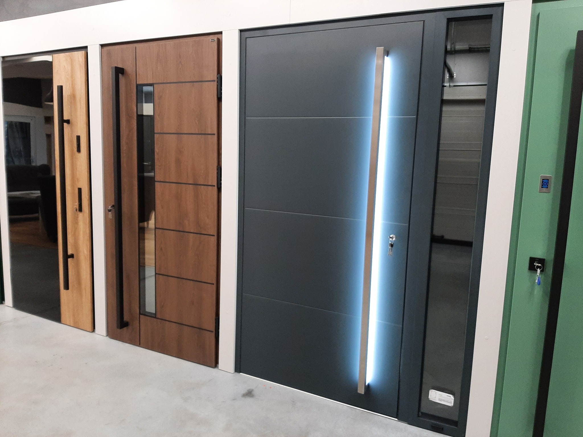 Wystawa trzech różnych modeli drzwi zewnętrznych: jasne drewnopodobne z pionowym przeszkleniem, ciemne drewnopodobne z poziomymi wstawkami i grafitowe z podświetlanym uchwytem, wszystkie z długimi...