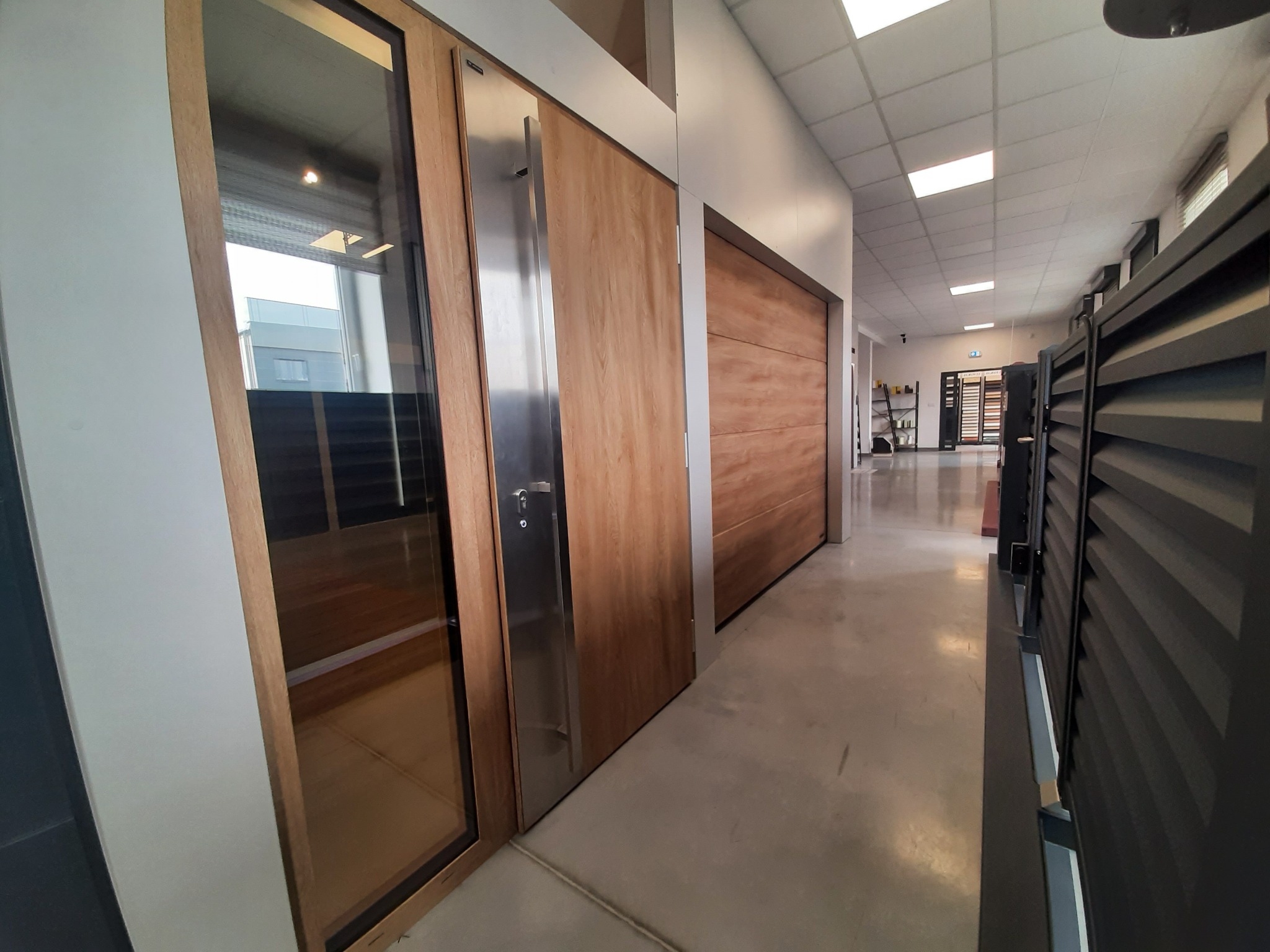 Ekspozycja w salonie: drewniane drzwi z dużym, pionowym, metalowym uchwytem oraz przeszklona witryna obok, na podłodze betonowa wylewka, w tle widoczne rolety.