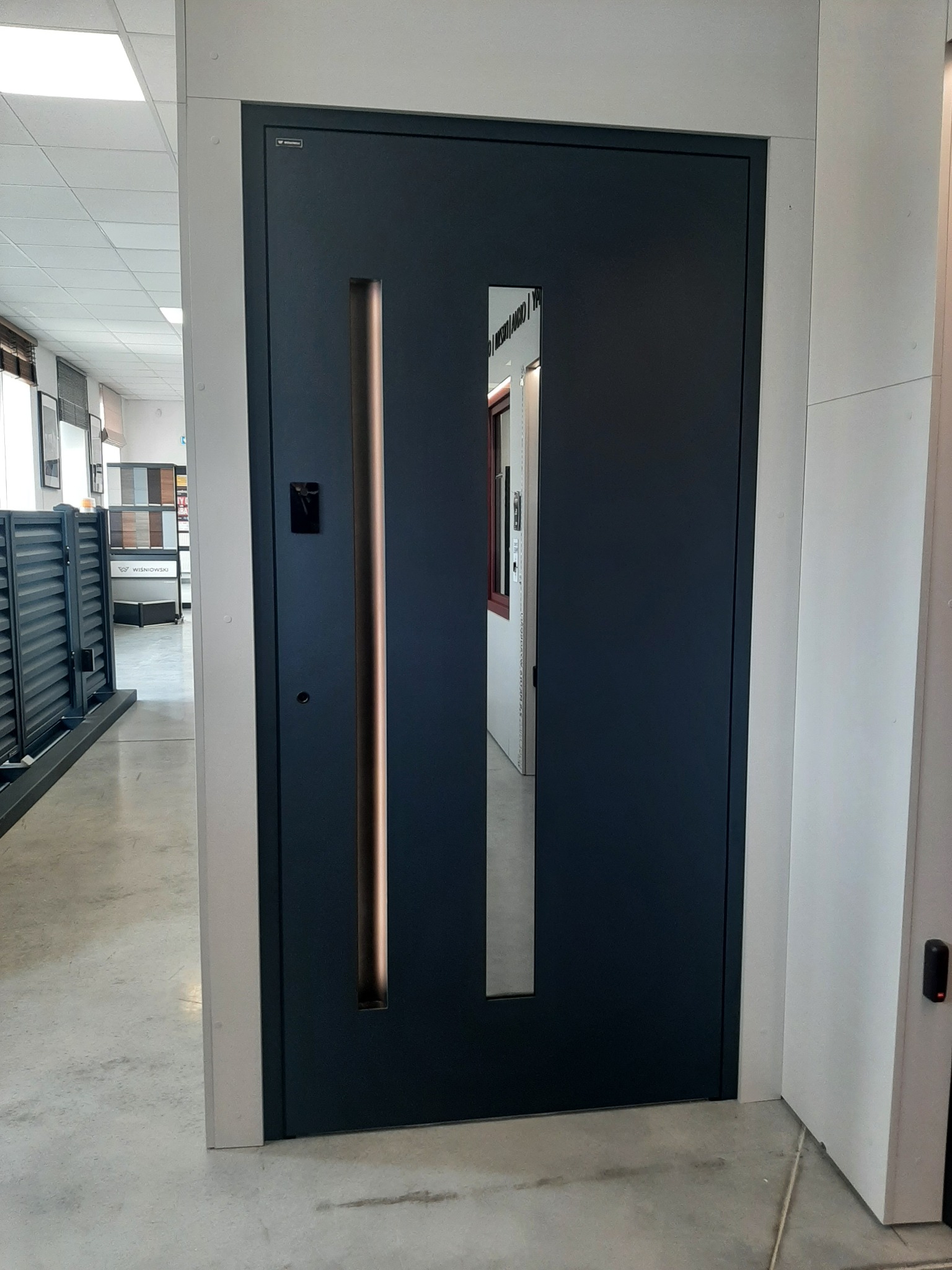 Ciemnogranatowe drzwi wejściowe z pionowym, podświetlanym panelem i prostokątnym wizjerem, prezentowane w salonie wystawowym.