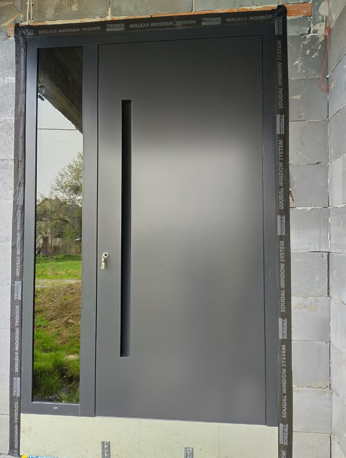 Nowoczesne, antracytowe drzwi wejściowe z przeszkleniem bocznym, zamontowane w surowym stanie, widoczne detale montażu i otoczenie budowy w Choczni.