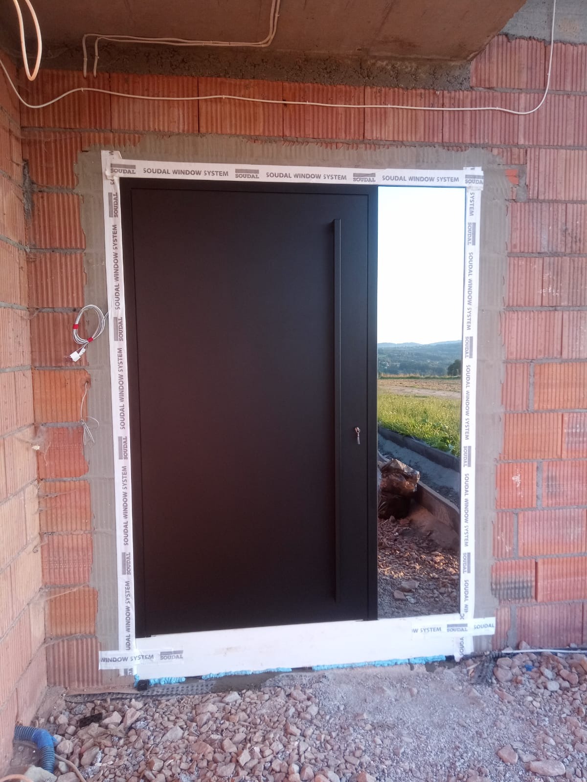 Montaż czarnych drzwi zewnętrznych w surowym murze z cegły, widoczne uszczelnienia Soudal Window System, otwarte drzwi ukazują krajobraz za domem.