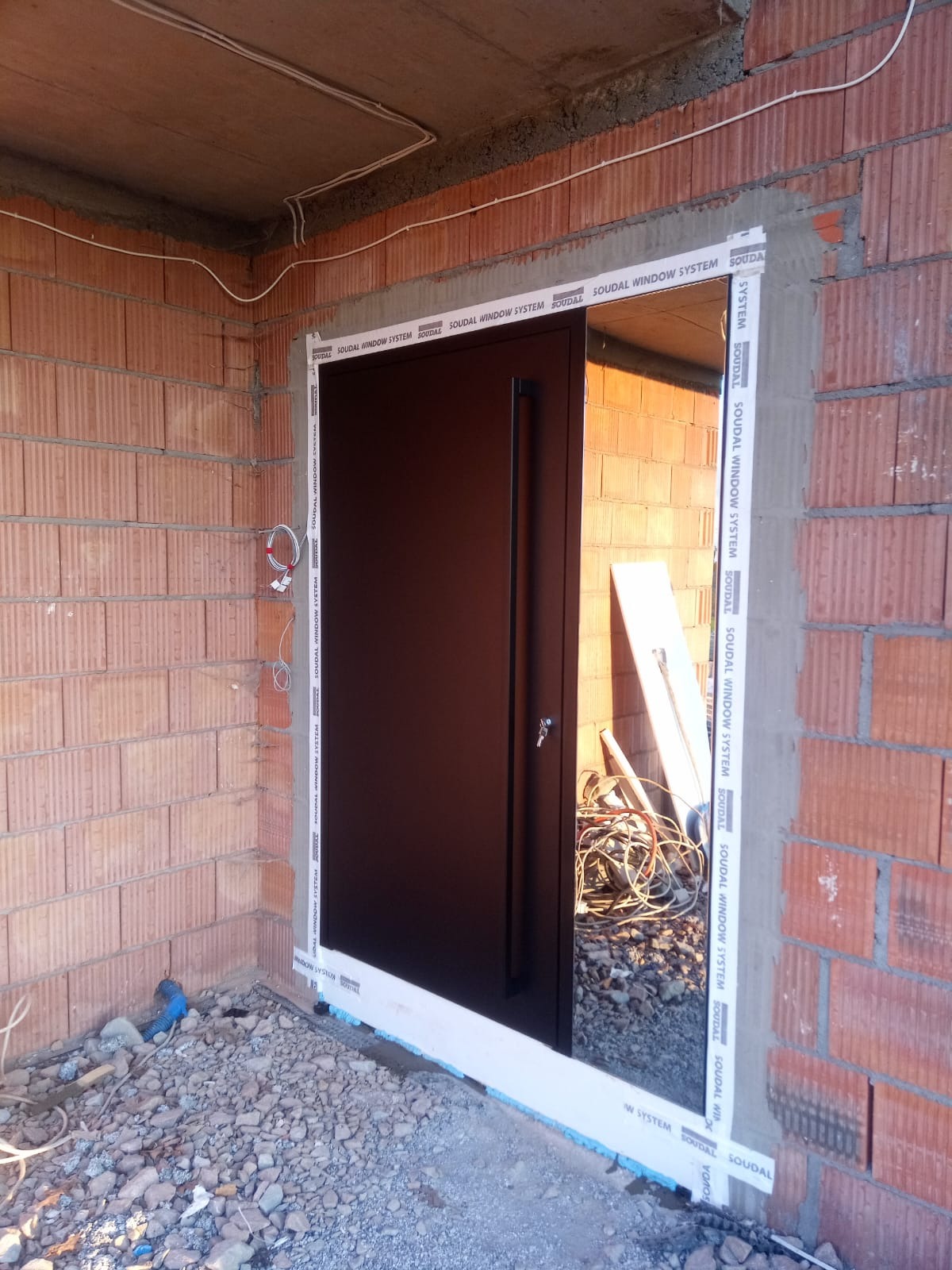 Montaż czarnych drzwi wejściowych w surowym stanie, widoczne cegły, przewody i materiały budowlane.