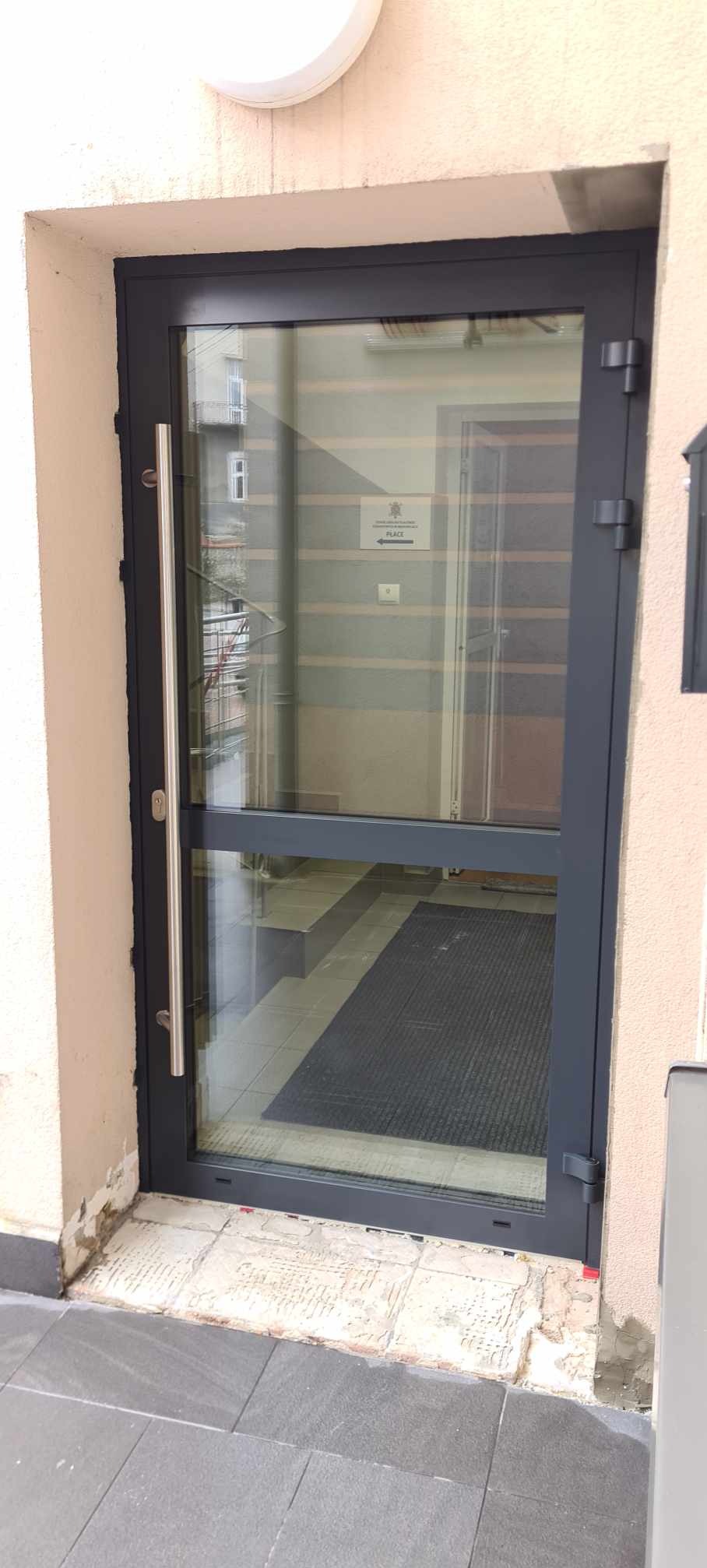 Szklane drzwi wejściowe z długim, pionowym uchwytem ze stali nierdzewnej, widoczny znak informacyjny wewnątrz budynku, ślady prac budowlanych przy progu.