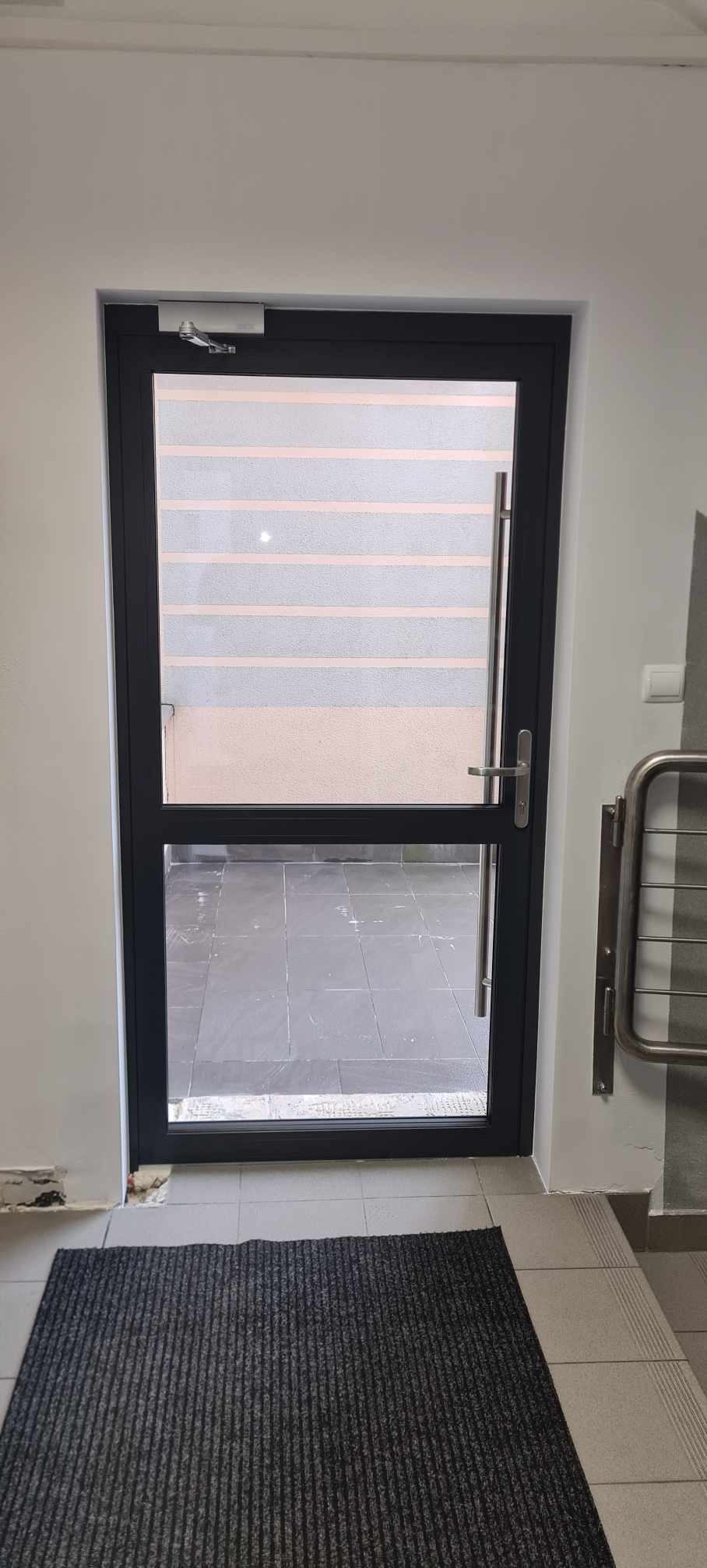 Ciemna rama przeszklonych drzwi zewnętrznych z samozamykaczem, widok na balkon z szarymi płytkami, wycieraczka przed wejściem.