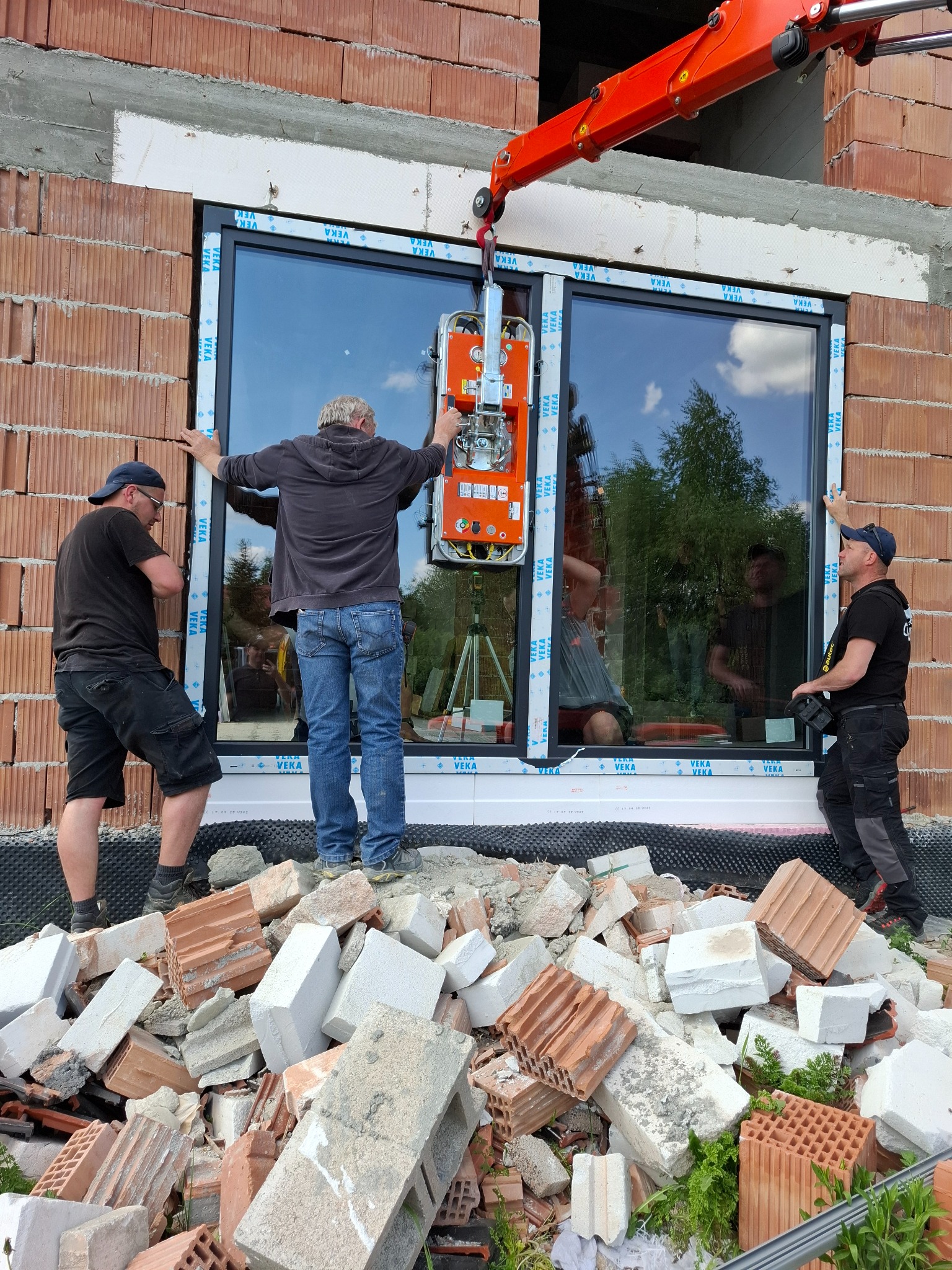 Montaż okna PCV z użyciem przyssawki i dźwigu na placu budowy. Trzech monterów stabilizuje ramę okienną marki VEKA. Widoczne gruz i cegły.