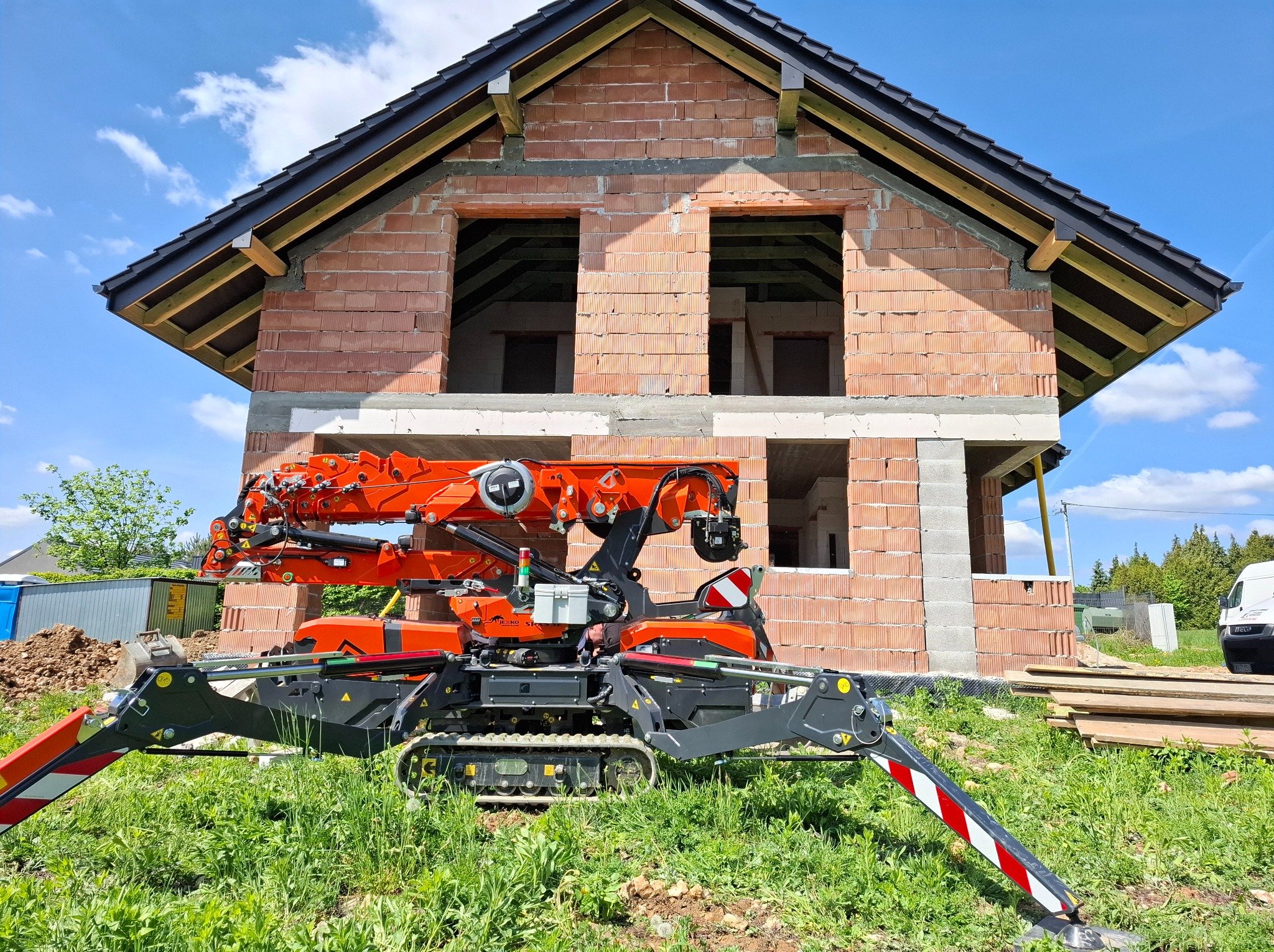 Mini żuraw pająk na tle surowego budynku z czerwonej cegły, z widocznym dachem i otworami okiennymi, na zielonej trawie w słoneczny dzień.