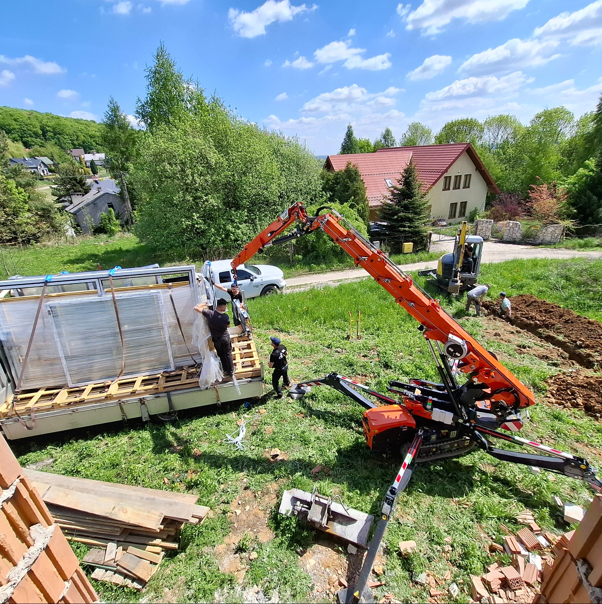 Rozładunek okien z użyciem dźwigu na budowie domu jednorodzinnego w słoneczny dzień. Widoczni pracownicy i wykop pod fundamenty.