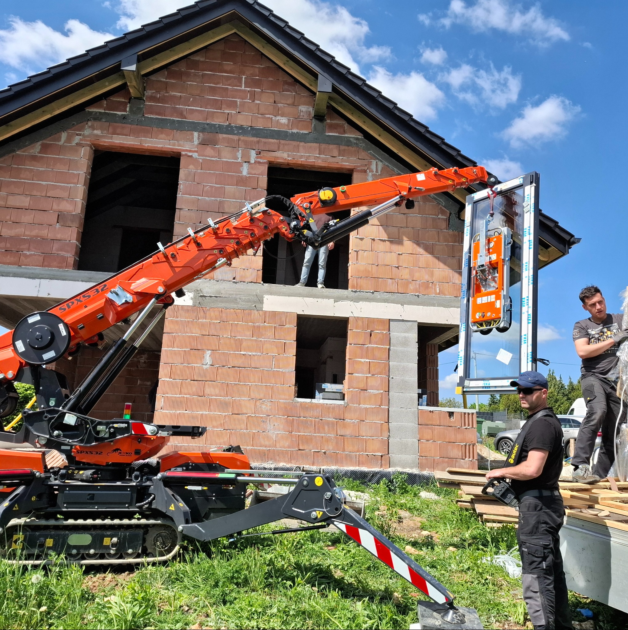 Montaż okna za pomocą minikoparki z przyssawkami na budowie domu z czerwonej cegły. Operator steruje maszyną, drugi pracownik pomaga w montażu.