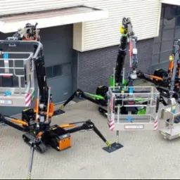 Dwa pająkowe podnośniki koszowe ustawione obok budynku o szarej fasadzie, jeden z koszem oznaczonym 'FAB 2', drugi z zielonymi elementami i koszem z napisem 'FERRARI'.