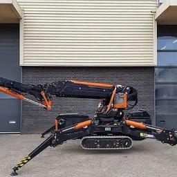 Megaton - Pająkowy minidźwig gąsienicowy BG Lift SAK 100 z wysuniętym ramieniem, zaparkowany na zewnątrz budynku z szarą bramą rolowaną i ceglaną ścianą.