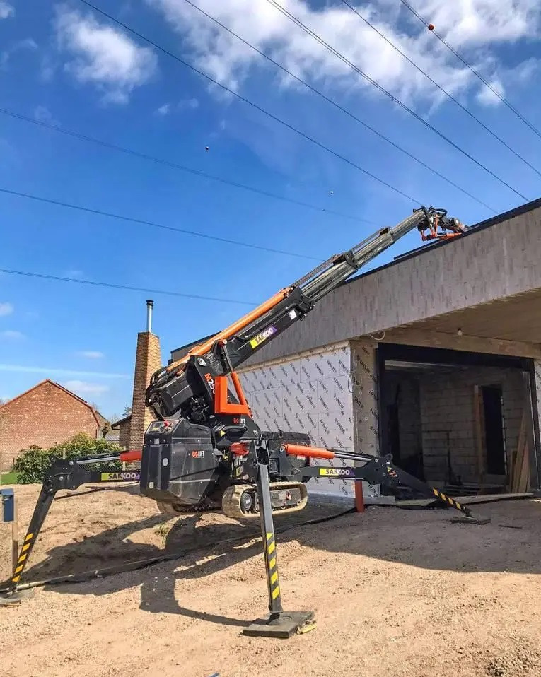 Czarny pająk-dźwig Sakoo M250 na gąsienicach, z wysuniętymi podporami, sięga pomarańczowym ramieniem do krawędzi dachu budynku w trakcie budowy, na tle błękitnego nieba z liniami energetycznymi.