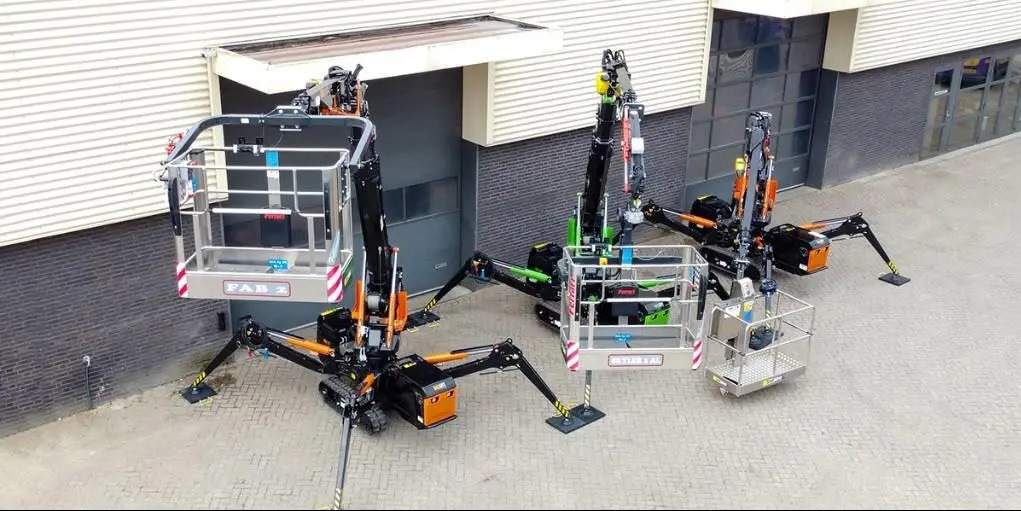 Dwa pająkowe podnośniki koszowe ustawione obok budynku o szarej fasadzie, jeden z koszem oznaczonym 'FAB 2', drugi z zielonymi elementami i koszem z napisem 'FERRARI'.