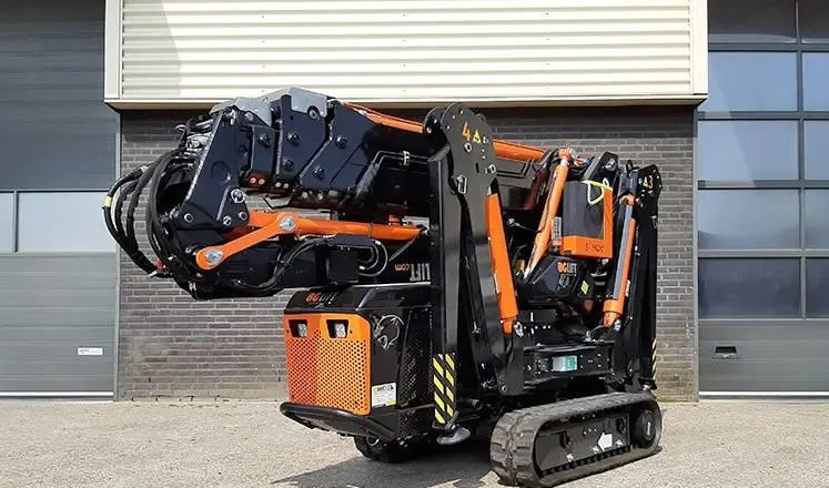 Kompaktowa gąsienicowa maszyna z ramieniem teleskopowym, czarno-pomarańczowa kolorystyka, na tle szarej bramy garażowej i ceglanej ściany.
