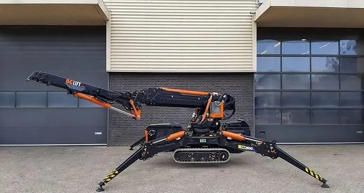 Pająkowy minidźwig gąsienicowy BG Lift SAK 100 z wysuniętym ramieniem, zaparkowany na zewnątrz budynku z szarą bramą rolowaną i ceglaną ścianą.