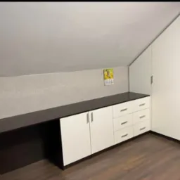 Funkcjonalna zabudowa pod skosem: biało-brązowe szafki i szuflady z ciemnym blatem, dopasowane do nietypowej przestrzeni poddasza. Na ścianie wisi kalendarz.