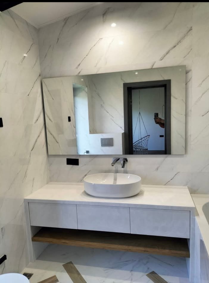 Nowoczesna łazienka z umywalką nablatową, szafką z drewnianą półką i dużym lustrem na marmurowej ścianie. W lustrze odbija się huśtawka.