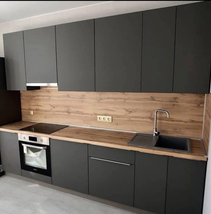 Nowoczesna kuchnia z szarymi matowymi frontami i drewnianym blatem. Widoczny piekarnik, płyta grzewcza i zlewozmywak. Minimalistyczny design z subtelnym oświetleniem.