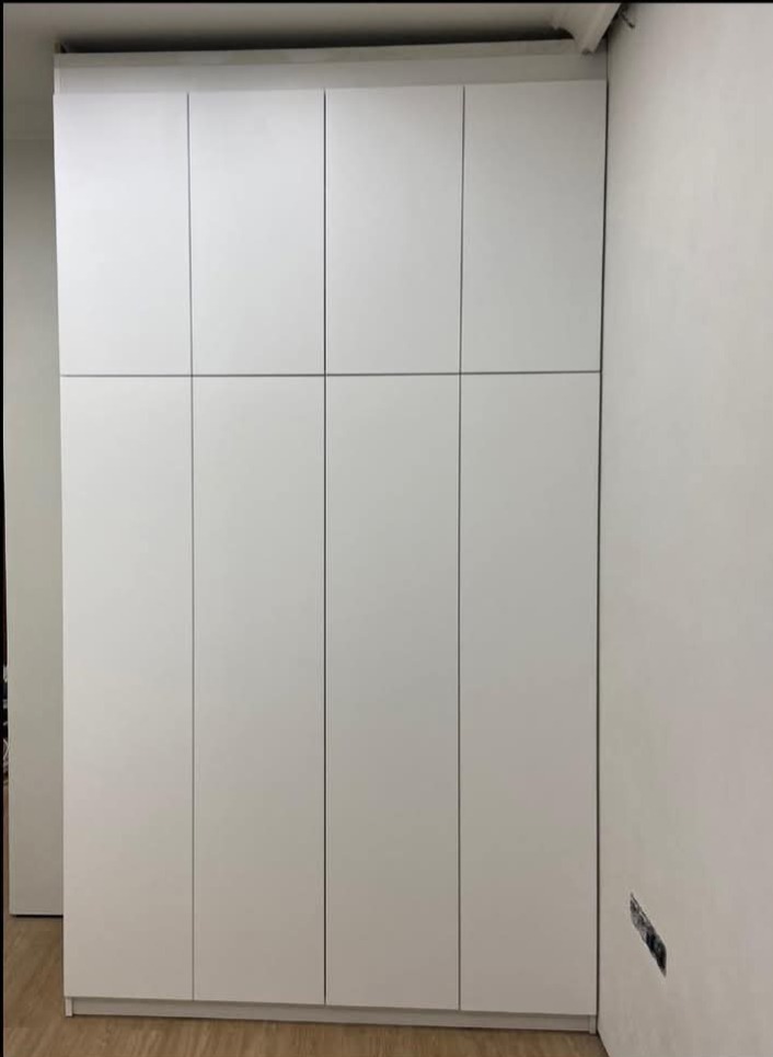 Biała, nowoczesna szafa wnękowa z ośmioma frontami, sięgająca sufitu, na tle jasnej ściany i podłogi. Widoczny fragment gniazdka elektrycznego w ścianie.