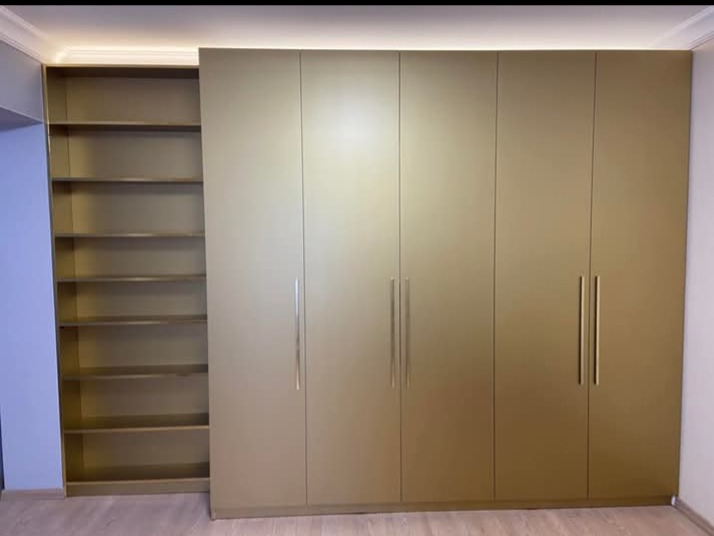 Złota szafa na wymiar z półkami w Nekli. Minimalistyczny design, długie uchwyty, oświetlenie LED nad szafą. Jasne wnętrze półek, podłoga imitująca drewno.