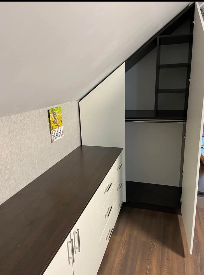 Szafa wnękowa na poddaszu ze skośnym sufitem, białe fronty, ciemny blat i wnętrze. Widoczny kalendarz na ścianie i drewniana podłoga.