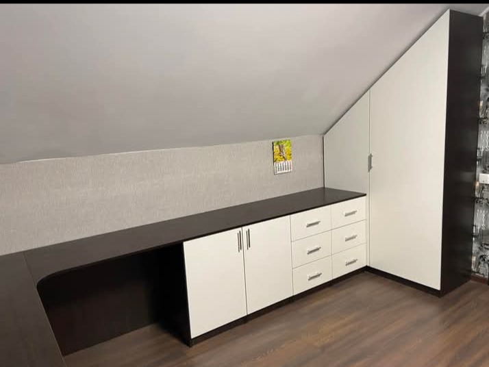 Funkcjonalna zabudowa pod skosem: biało-brązowe szafki i szuflady z ciemnym blatem, dopasowane do nietypowej przestrzeni poddasza. Na ścianie wisi kalendarz.