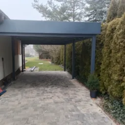 Nowoczesna, metalowa konstrukcja zadaszenia nad podjazdem, przylegająca do budynku, z widocznymi elementami wykończenia i narzędziami w tle, otoczona tujami.