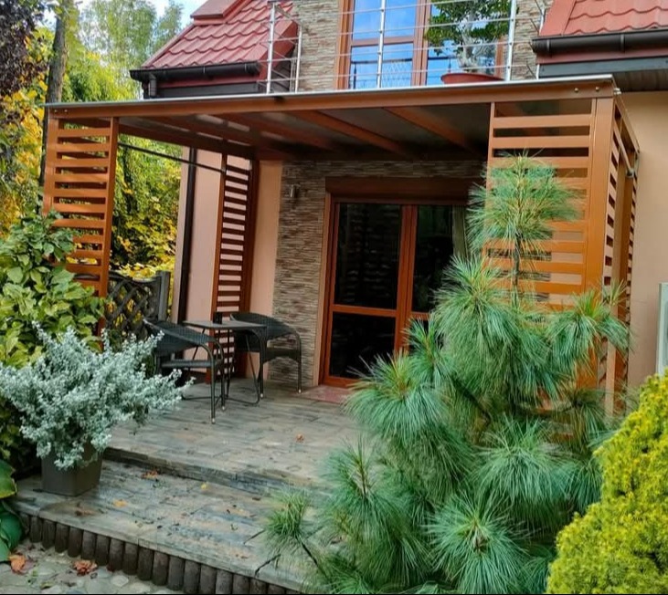 Zadaszona przestrzeń tarasowa z drewnianą konstrukcją, ażurowymi ściankami i meblami ogrodowymi, otoczona zielenią.