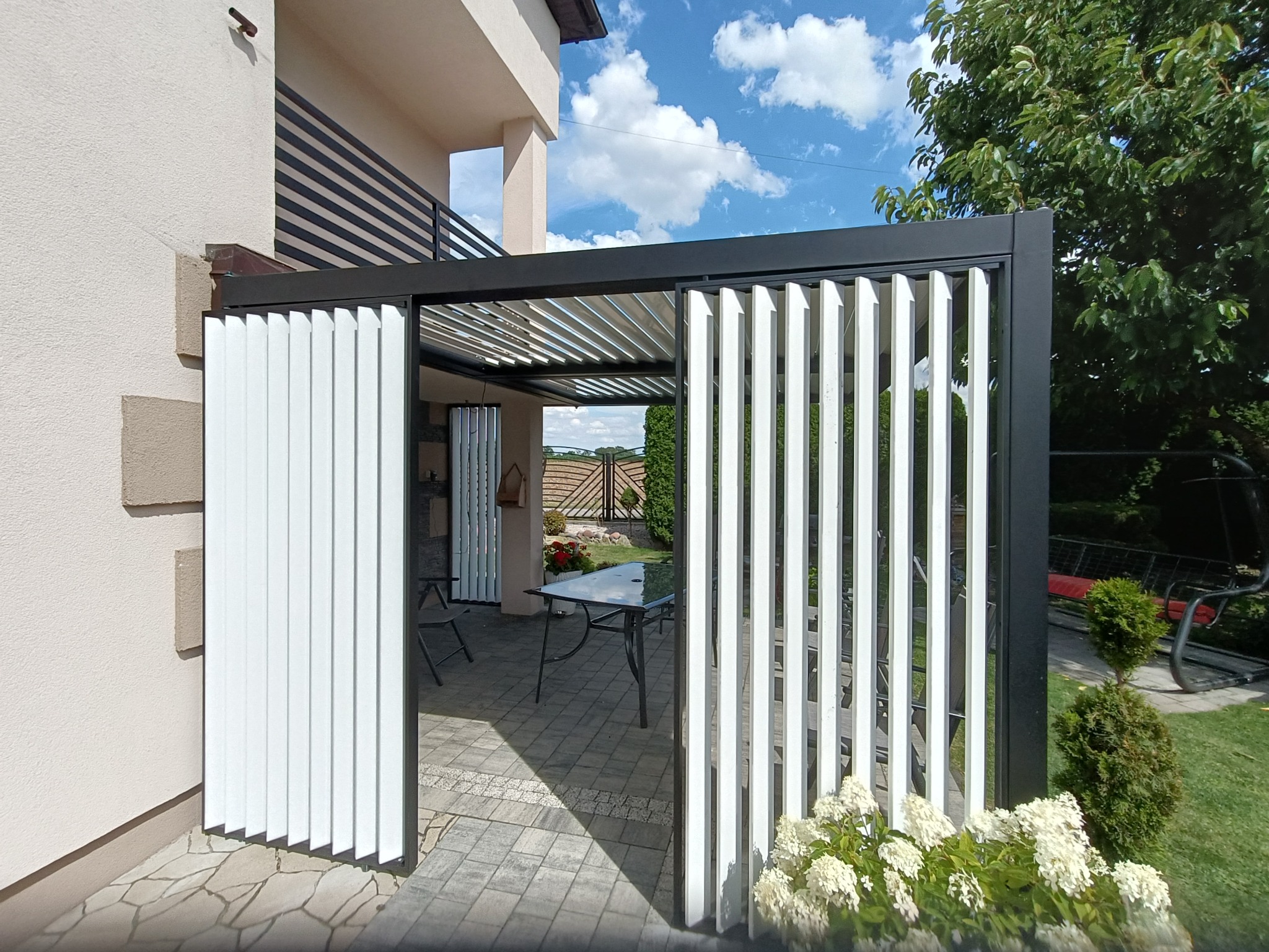 Nowoczesna pergola z ruchomymi, białymi panelami, przylegająca do budynku z balkonem, widok na ogród i błękitne niebo.