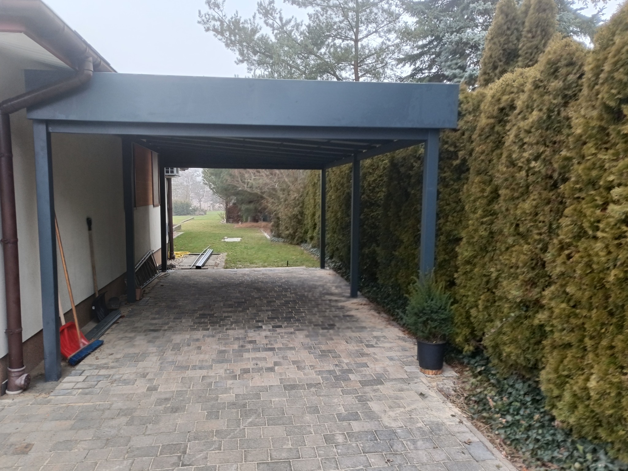 Nowoczesna, metalowa konstrukcja zadaszenia nad podjazdem, przylegająca do budynku, z widocznymi elementami wykończenia i narzędziami w tle, otoczona tujami.