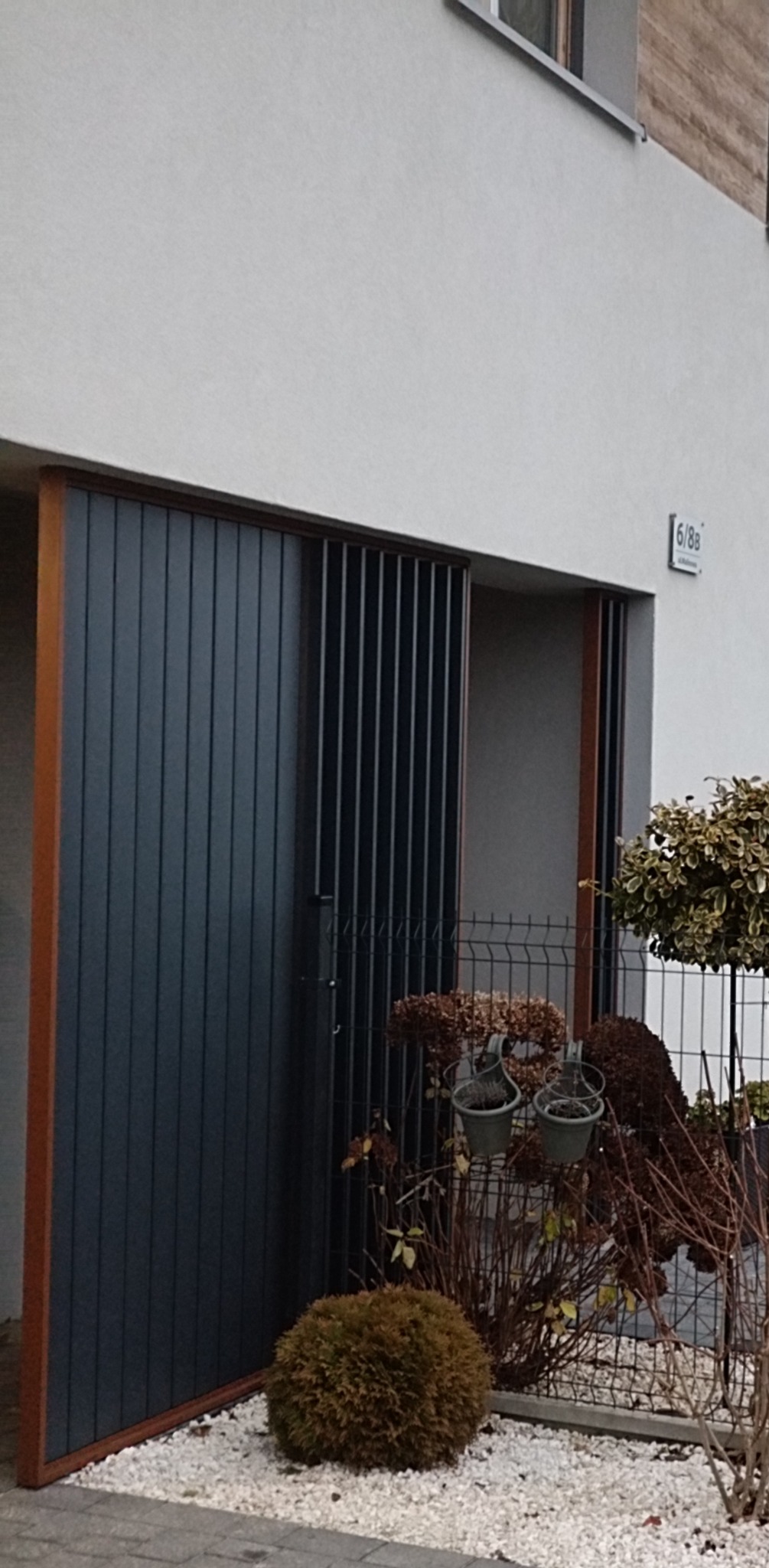 Nowoczesna brama wjazdowa z pionowych paneli, z częściowo otwartym ażurowym segmentem, widoczna z poziomu gruntu, z roślinami i białym żwirem w tle.