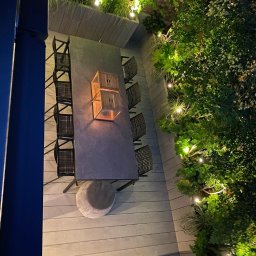 Marcin Kłosowski - Zewnętrzna jadalnia z długim stołem, krzesłami z plecionki, szarym pufem i oświetleniem dekoracyjnym wśród zieleni.