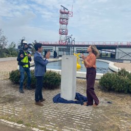 Marcin Kłosowski - Uroczyste odsłonięcie betonowego obelisku na zewnątrz budynku z wieżą, transmitowane przez kamerzystę, z udziałem kobiety w brązowej bluzce i mężczyzny w niebieskiej marynarce, oboje klaszczący.