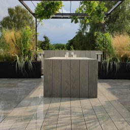 Marcin Kłosowski - Nowoczesny taras z szarych desek kompozytowych i płytek, czarne donice z ozdobnymi trawami, pergola z zielenią, minimalistyczny stół z dwoma szklankami wody, po deszczu.