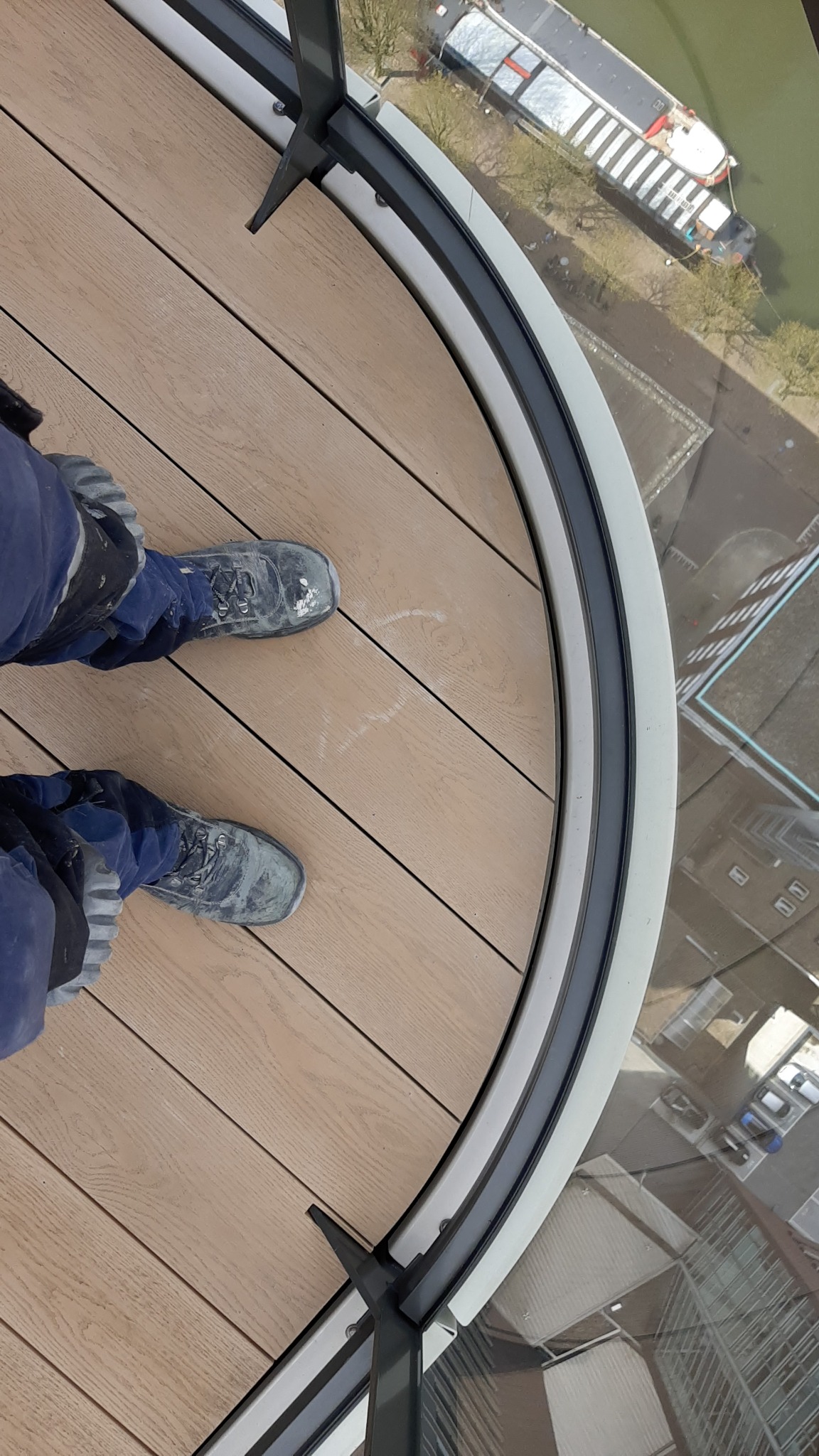 Widok z góry na fragment zabudowanego balkonu z drewnianą podłogą i widocznymi butami roboczymi pracownika, zza szyby widać budynki i rzekę z barkami.