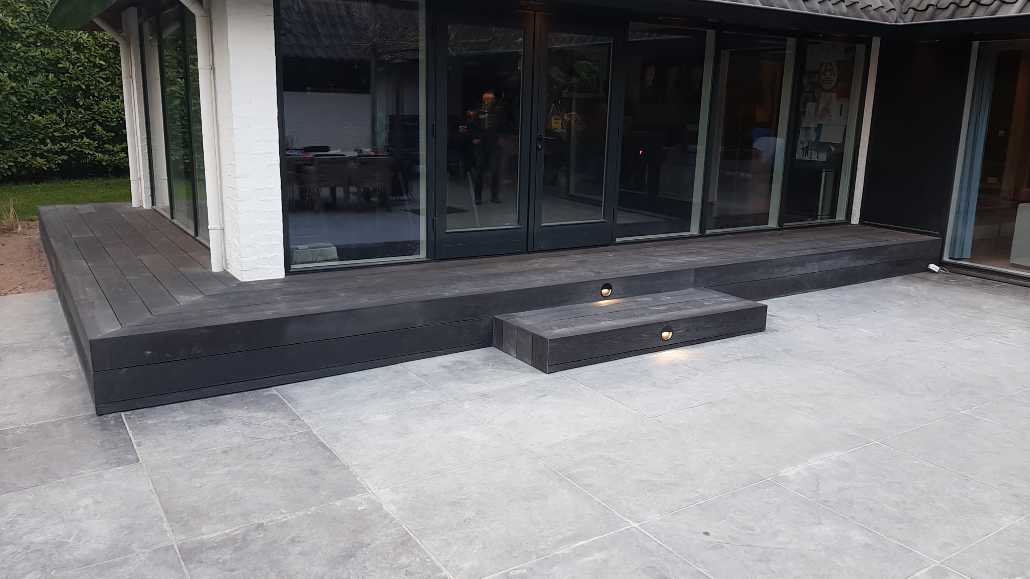 Nowoczesny taras z ciemnego drewna kompozytowego, z podświetlanymi schodkami, wyłożony jasnoszarą kostką brukową.