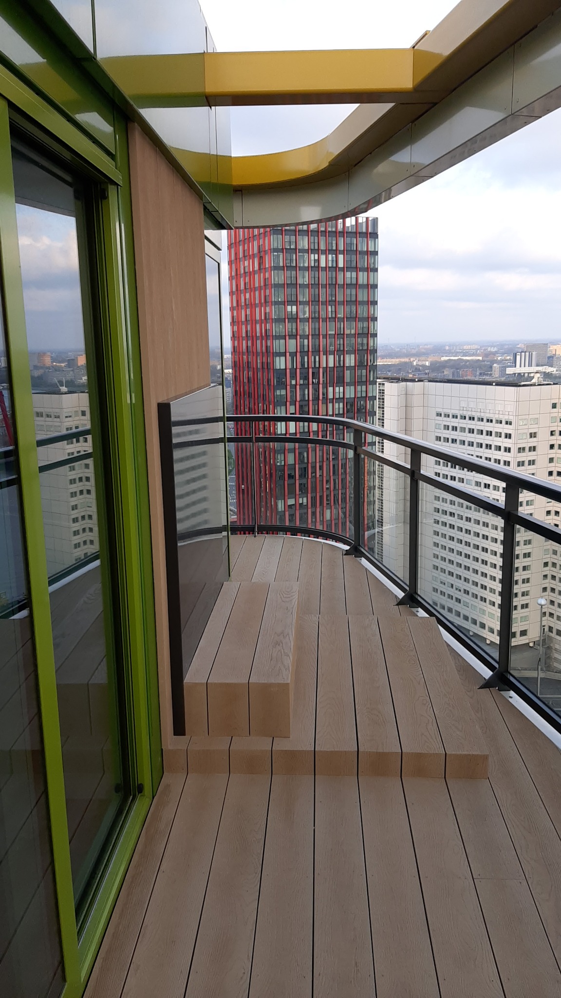 Nowoczesny balkon z drewnianą podłogą i szklaną balustradą w Gdańsku, z widokiem na panoramę miasta i charakterystyczny czerwony wieżowiec.