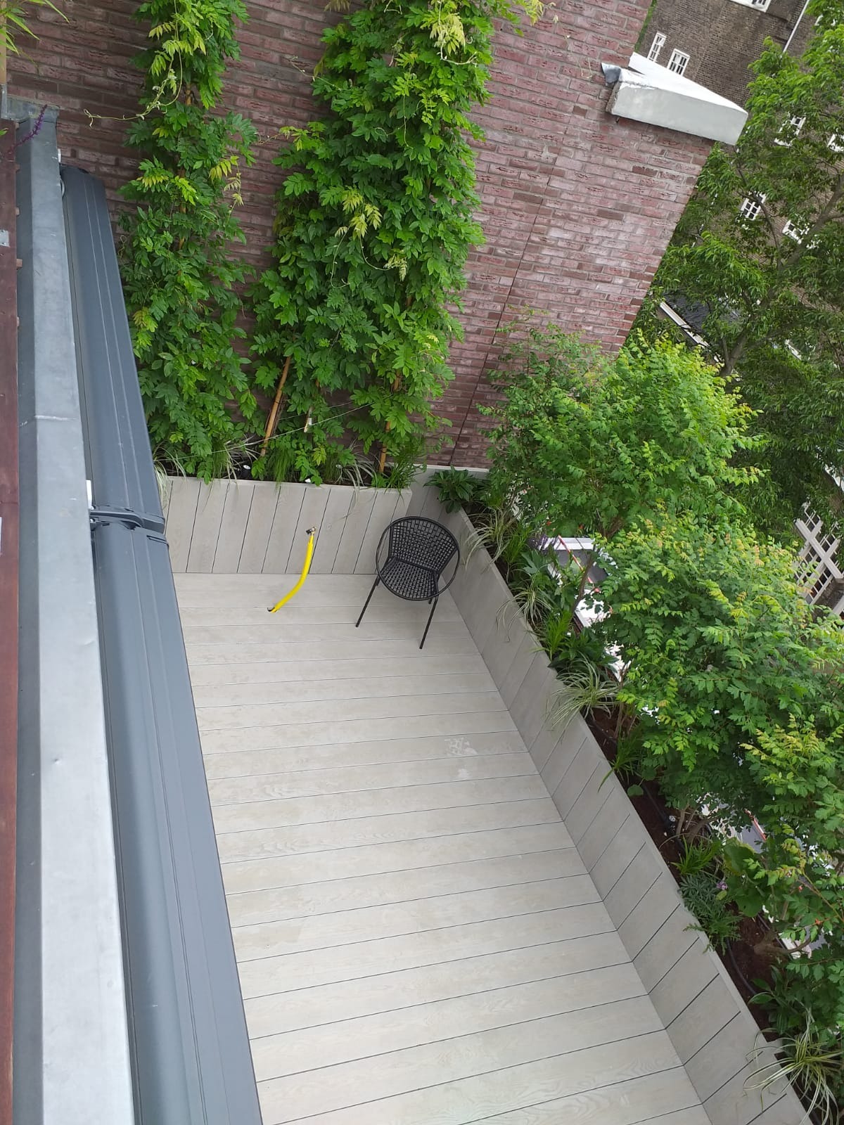 Widok z góry na zagospodarowany balkon z drewnianą podłogą, metalowym krzesłem i roślinami pnącymi się po ceglanej ścianie budynku.