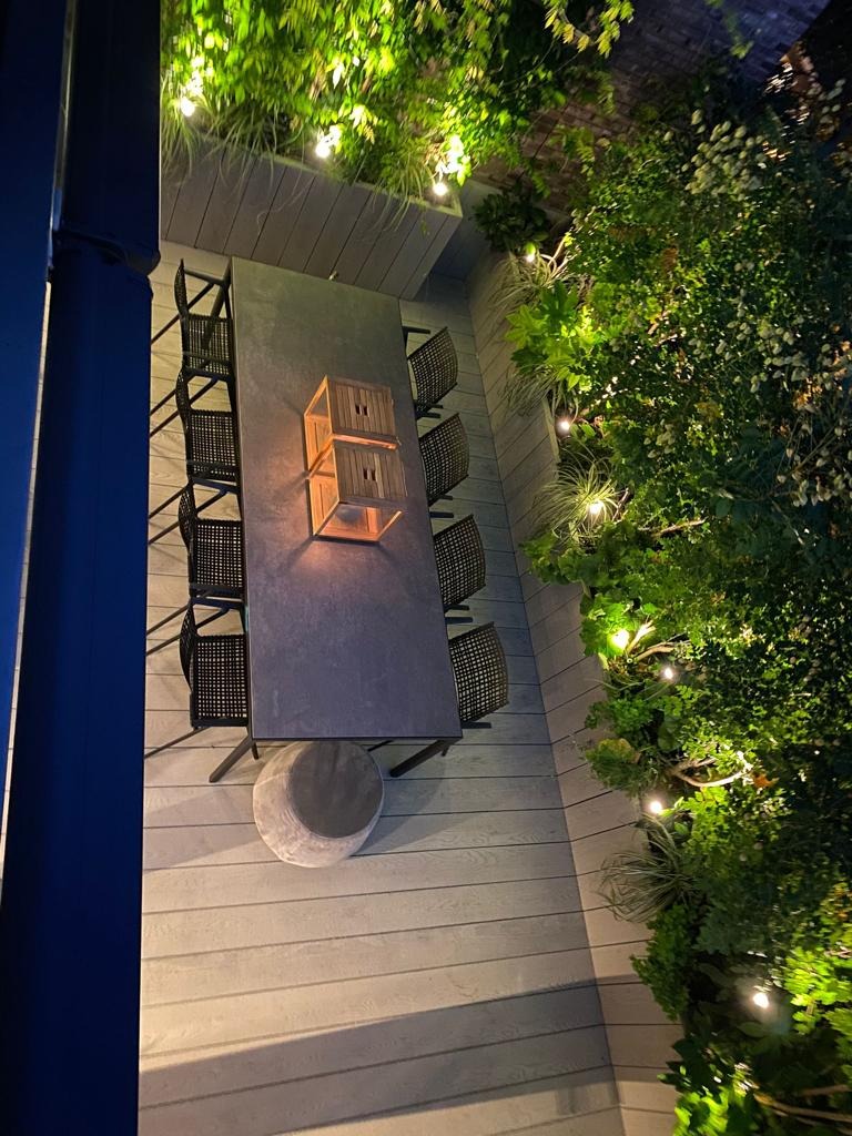 Zewnętrzna jadalnia z długim stołem, krzesłami z plecionki, szarym pufem i oświetleniem dekoracyjnym wśród zieleni.