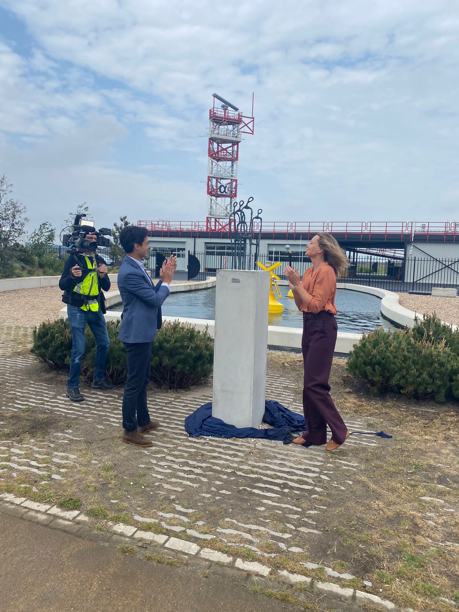 Uroczyste odsłonięcie betonowego obelisku na zewnątrz budynku z wieżą, transmitowane przez kamerzystę, z udziałem kobiety w brązowej bluzce i mężczyzny w niebieskiej marynarce, oboje klaszczący.