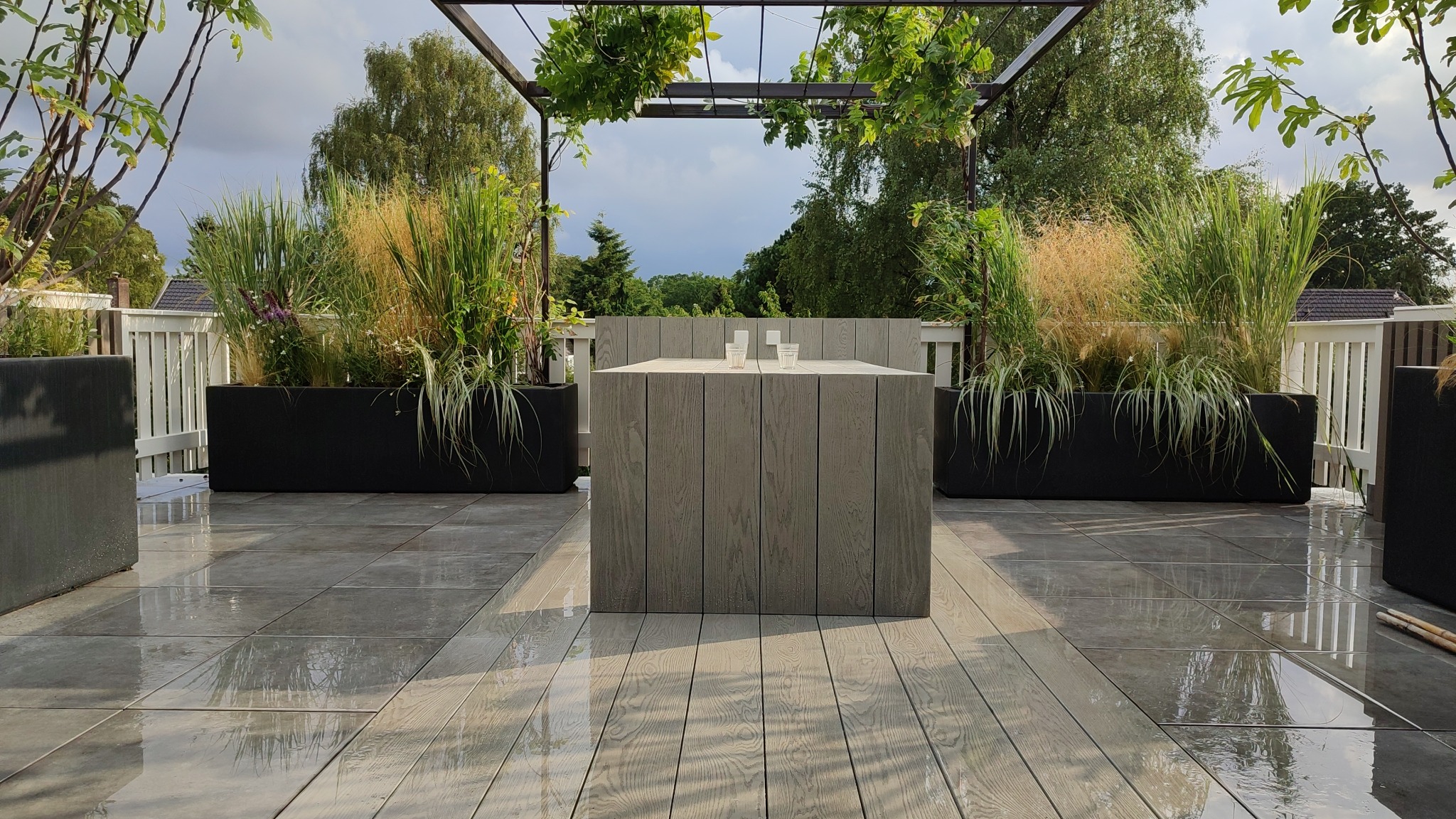 Nowoczesny taras z szarych desek kompozytowych i płytek, czarne donice z ozdobnymi trawami, pergola z zielenią, minimalistyczny stół z dwoma szklankami wody, po deszczu.