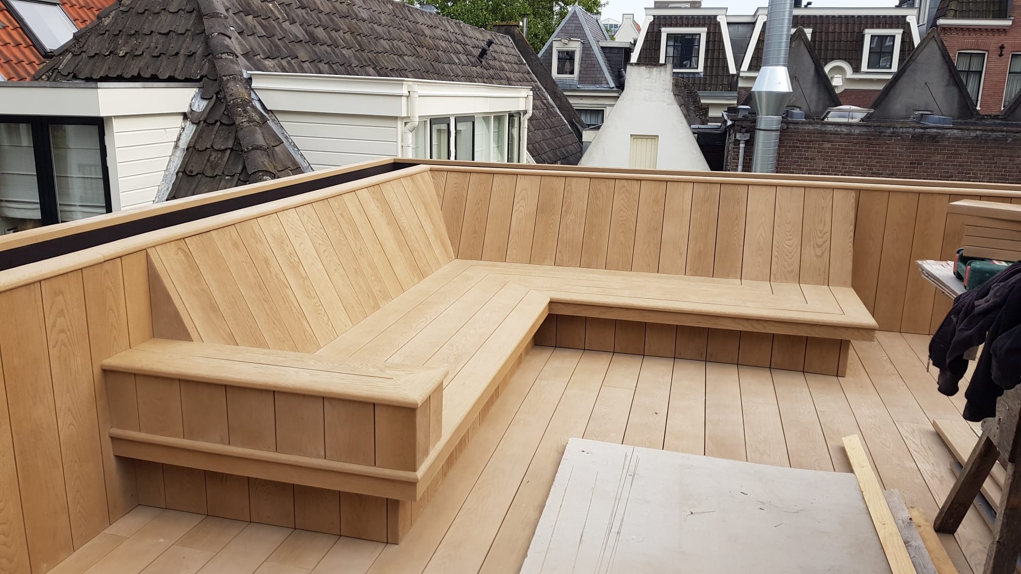 Drewniany taras na dachu z zabudowaną ławką w kształcie litery L, widok na dachy sąsiednich budynków.