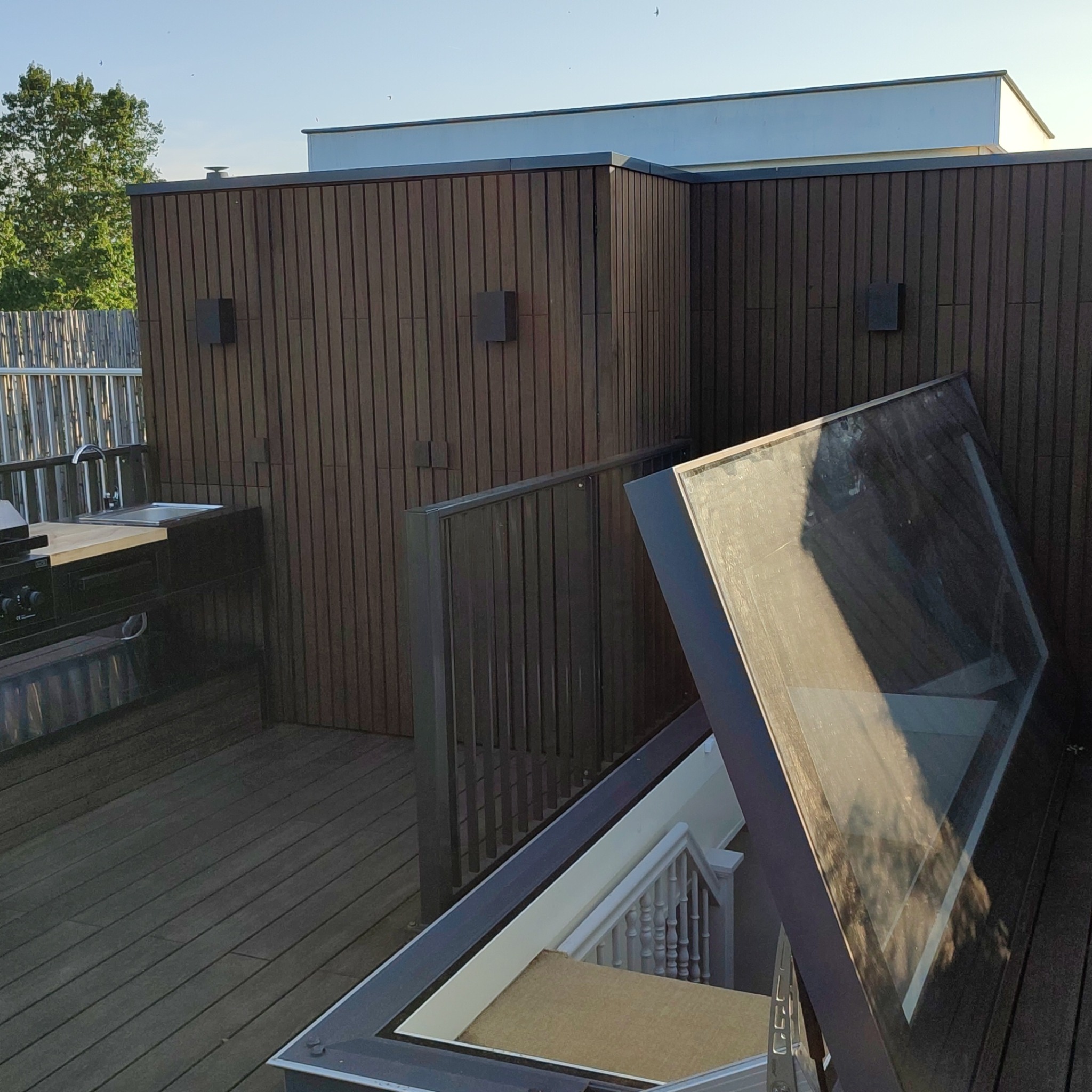 Drewniany taras na dachu z zabudową z pionowych paneli, zewnętrzną kuchnią z grillem i otwartym zejściem po schodach zabezpieczonym szklaną klapą.