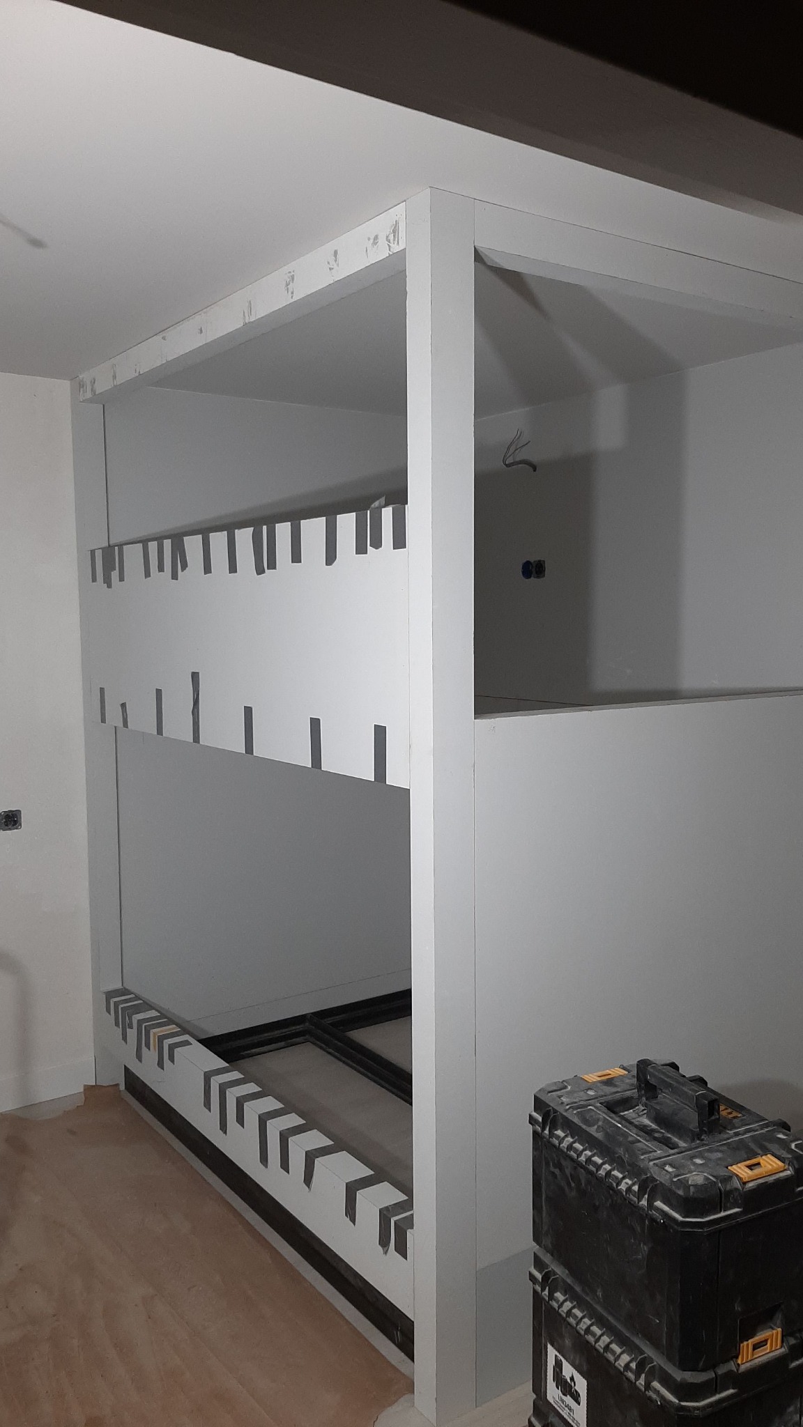 Wnętrze w trakcie tworzenia zabudowy z płyt kartonowo-gipsowych, widoczne fragmenty konstrukcji przytwierdzone taśmą, skrzynka z narzędziami w rogu pomieszczenia.