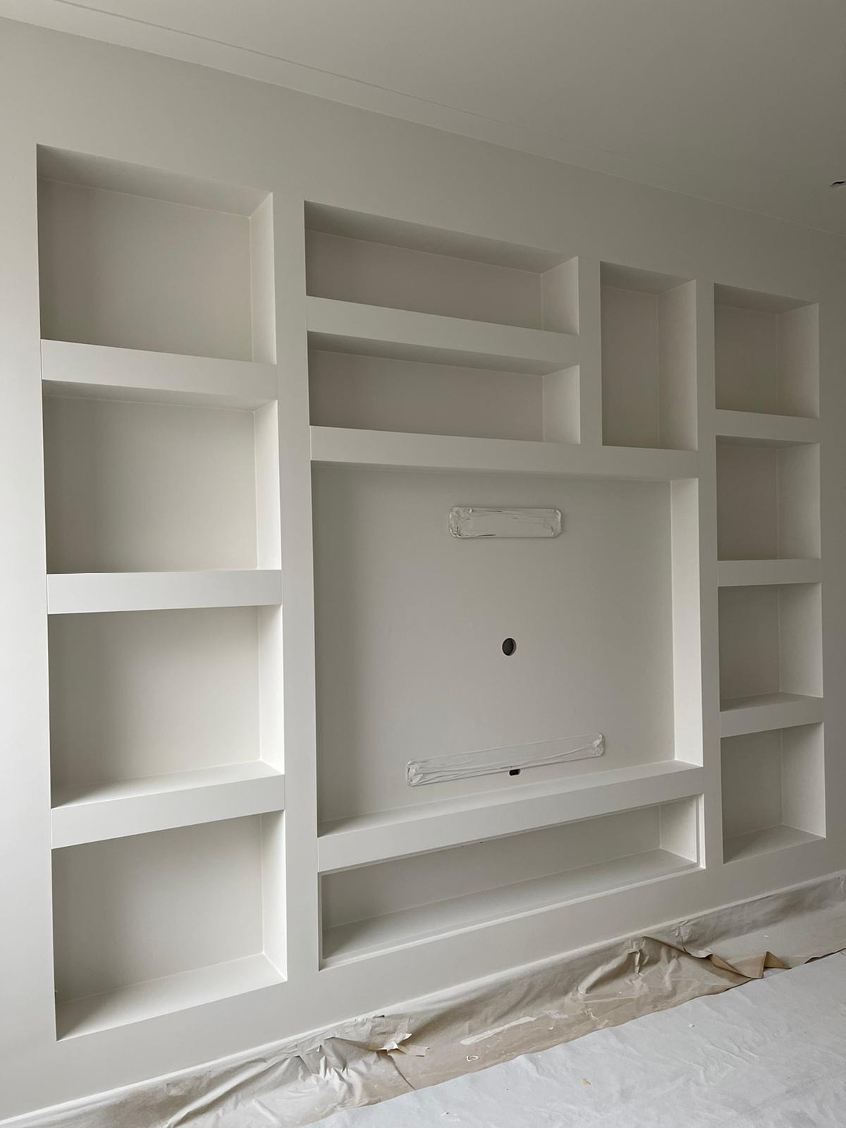 Biała zabudowa ściany z wnękami i półkami, przygotowana do montażu telewizora, zabezpieczona folią malarską na podłodze.